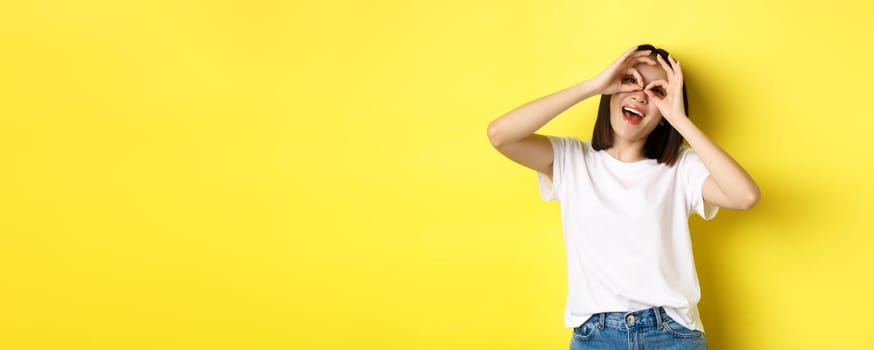 Funny asian girl looking through hand binoculars and smiling, stading over yellow background.