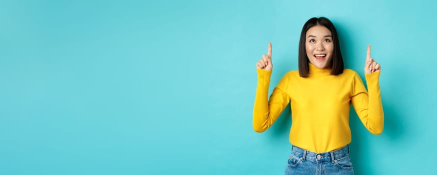 Shopping concept. Beautiful asian woman in yellow sweater pointing fingers up at logo, looking at promotion with happy smile, standing over blue background.