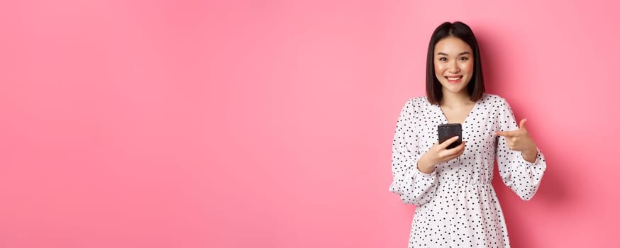 Beautiful young asian woman in romantic dress using smartphone, smiling and pointing at mobile phone, standing over pink background.