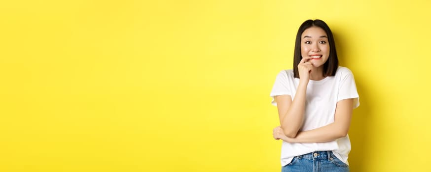 Portrait of cheerful asian girl wants something, looking with desire and temptation at camera, smiling at camera, standing ovr yellow background.