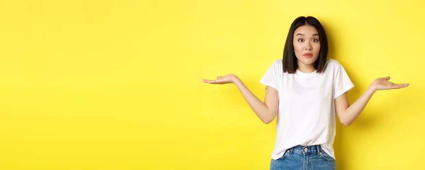 Clueless asian girl shrugging shoulders, spread out hands and looking confused, know nothing, standing over yellow background.