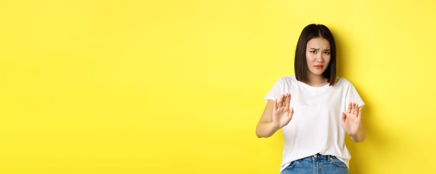 Please no. Asian woman, victim of assualt or domestic abuse, pleading with hands raised up in defense, frowning sad, begging stop, standing over yellow background.