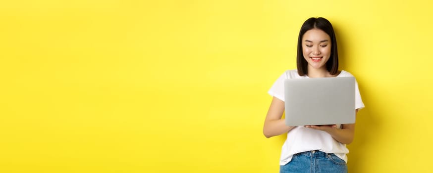 Beautiful asian woman working on laptop, looking happy at screen, standing over yellow background.