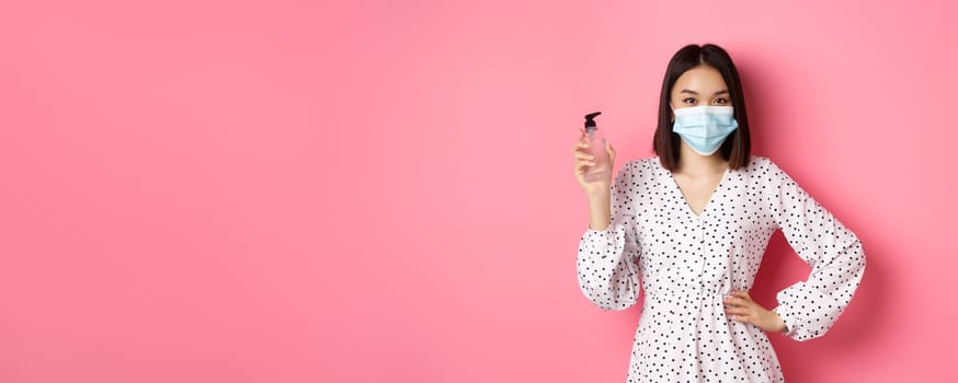 Covid-19, pandemic and lifestyle concept. Beautiful korean woman in dress and medical mask showing hand sanitizer, recommending antiseptic, standing over pink background.