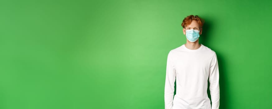 Covid-19, social distancing and lifestyle concept. Young redhead man wearing face mask during coronavirus pandemic, standing over green background.