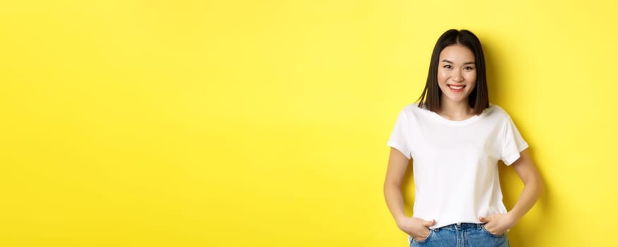 Beauty and fashion concept. Stylish asian girl in white t-shirt, smiling happy and standing over yellow background.