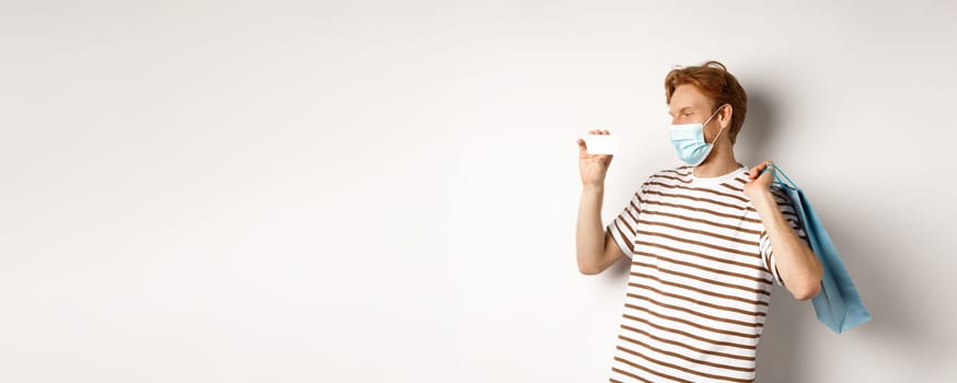 Concept of covid-19 and shopping. Happy young shopper in face mask holding paper bag and showing plastic credit card, buying with discounts, white background.