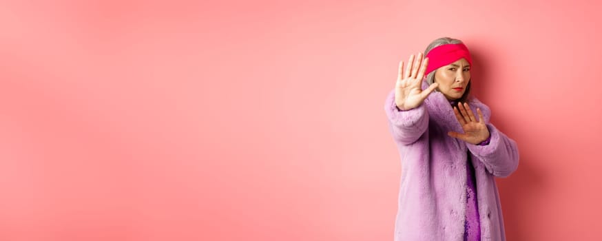 Asian senior female model in stylish purple winter coat standing in victim pose, extending hand in stop gesture and pleading for mercy, standing against pink background.