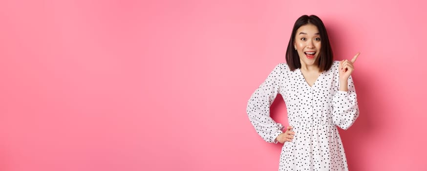 Excited pretty asian girl pointing at upper right corner, gasping amazed and looking at camera, discuss store promo offer, standing over pink background.