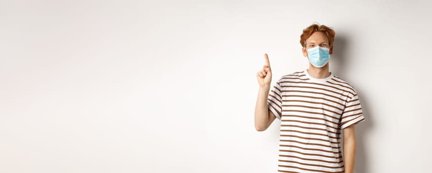 Covid, virus and social distancing concept. Young male student with red hair, using face mask from coronavirus, pointing finger up and smiling, white background.