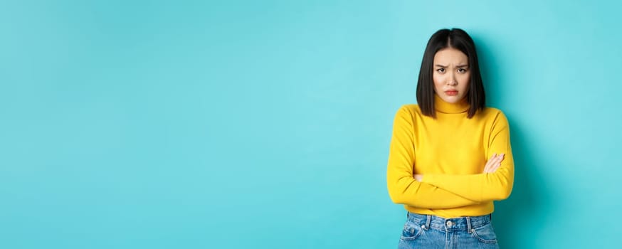 Image of asian girlfriend feeling offended, cross arms on chest and sulking, staring at camera insulted with something, standing over blue background.
