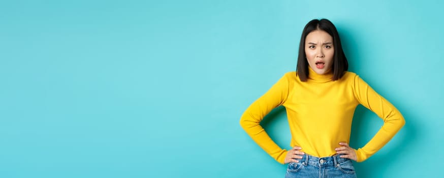 Portrait of angry and confused asian woman staring frustrated at camera, standing against blue background.