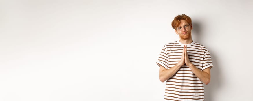 Silly redhead man asking for help, holding hands in pleading gesture and pucker lips, say please while begging you, standing over white background.