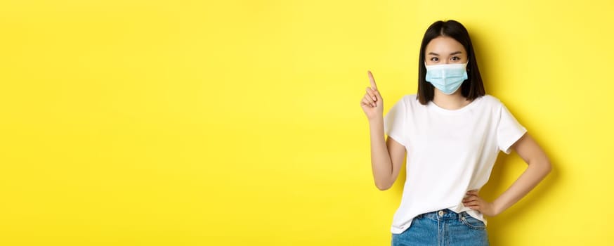 Covid, health care and pandemic concept. Asian female model in medical mask and white t-shirt pointing finger at upper left corner logo, showing promotion, yellow background.