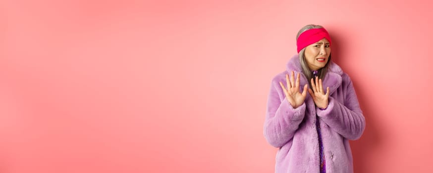 Portrait of stylish asian senior woman in purple winter coat, cringe from something disgusting, frowning and turn away awkward, showing stop gesture, refusing on pink background.