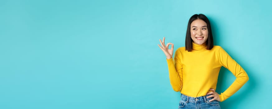 Impressed asian woman in yellow sweater, looking left amazed and showing OK sign in approval, standing over blue background.