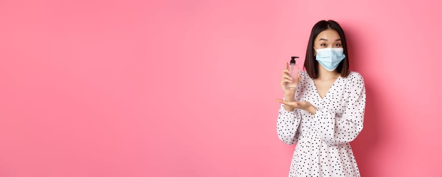 Covid-19, pandemic and lifestyle concept. Beautiful korean woman in dress and medical mask showing hand sanitizer, recommending antiseptic, standing over pink background.