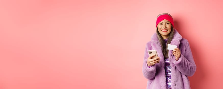 Online shopping and fashion concept. Smiling middle-aged woman in stylish fur coat using mobile phone and plastic credit card, standing happy on pink background.