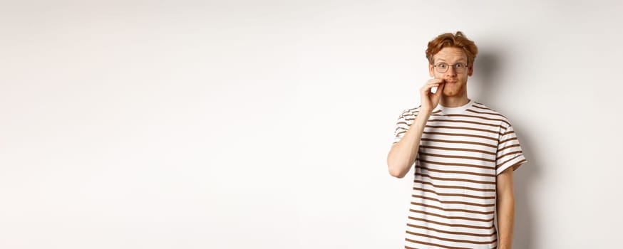 Young guy with ginger hair and glasses zipping mouth, showing lips seal gesture and smiling, keeping a secret, standing over white background.