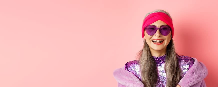 Fashion. Close up of stylish senior woman smiling happy at camera, wearing sunglasses and disco headband, pink background.