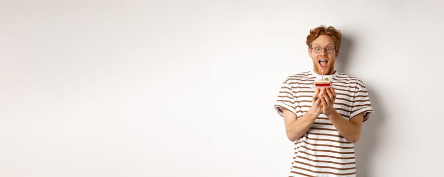 Holidays and celebration concept. Funny redhead guy blowing candle on birthday cake, smiling and making wish, white background.