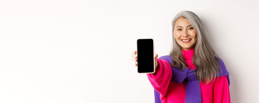 Online shopping. Close up of stylish asian senior woman extending hand with mobile phone, showing blank smartphone screen and smiling, standing over white background.