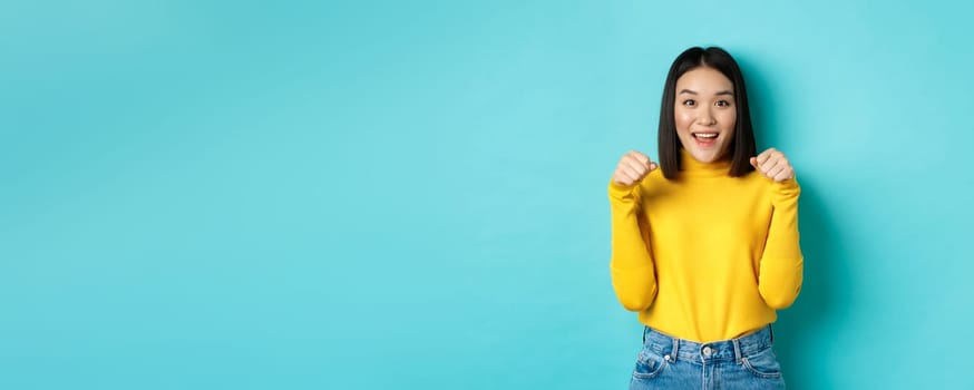 Beauty and fashion concept. Beautiful asian woman in yellow pullover raise hands as if holding logo, standing over blue background.