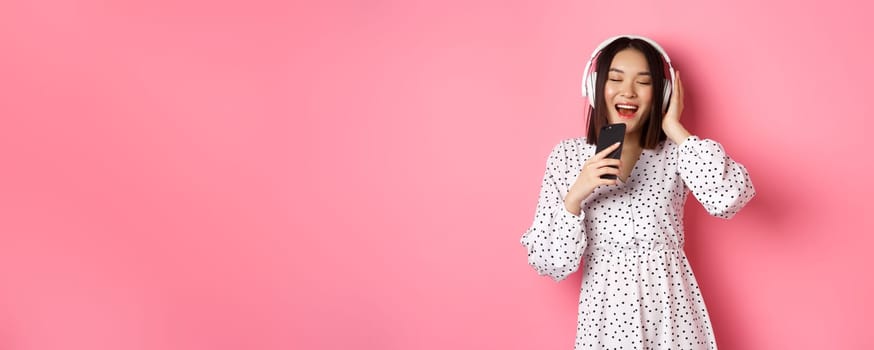 Cute asian woman playing karaoke app, singing in mobile phone and using headphones, standing in dress over pink background.