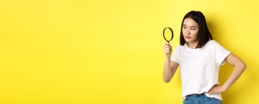 Asian woman detective looking through magnifying glass with intrigued look, found clues, standing over yellow background.