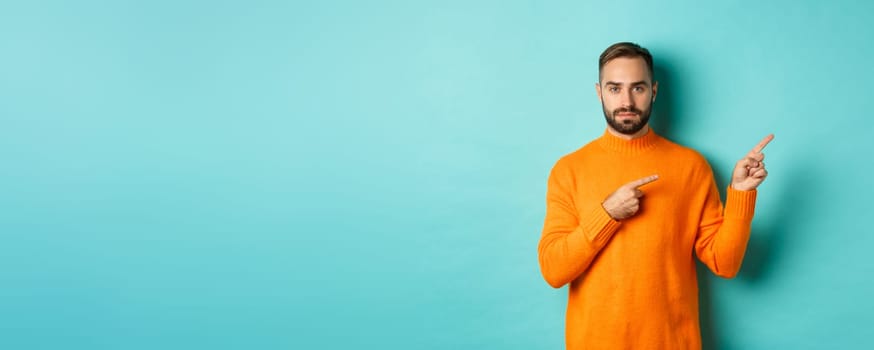 Handsome and serious bearded man in orange sweater pointing right, showing advertisement or logo, standing over turquoise background.