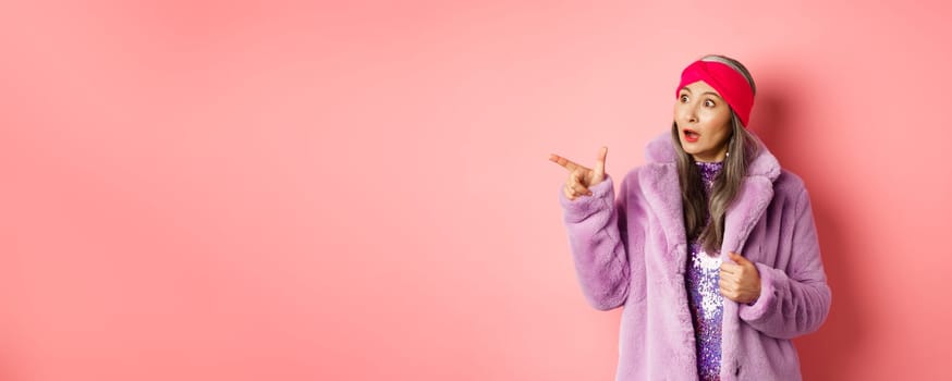 Fashion and shopping concept. Trendy elderly asian female in fake fur and glittering dress pointing, looking left with amazement, checking out offer, pink background.