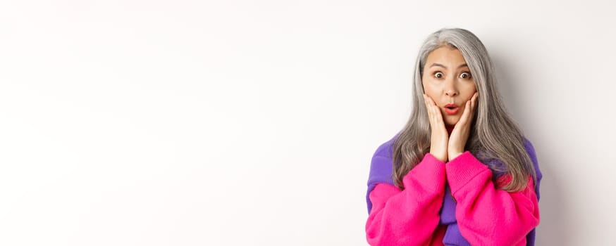Close up of fashionable asian senior woman with grey hair looking surprised, saying wow and staring at camera amazed, standing over white background.