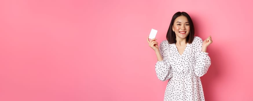 Shopping concept. Pretty asian woman holding bank credit card and smiling, standing over pink background.