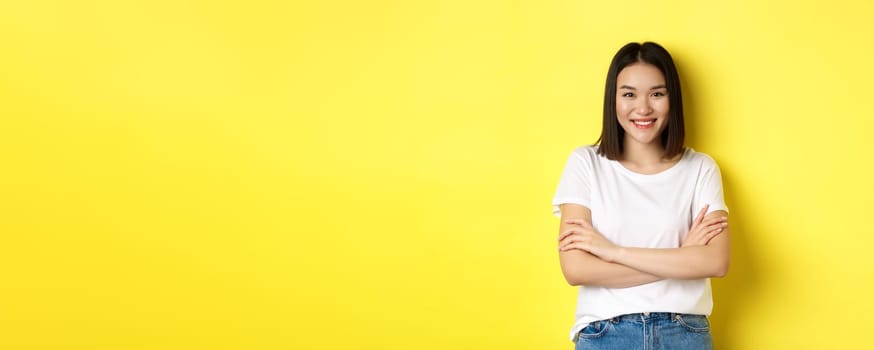 Confident and stylish asian woman cross arms on chest and smiling, standing over yellow background.