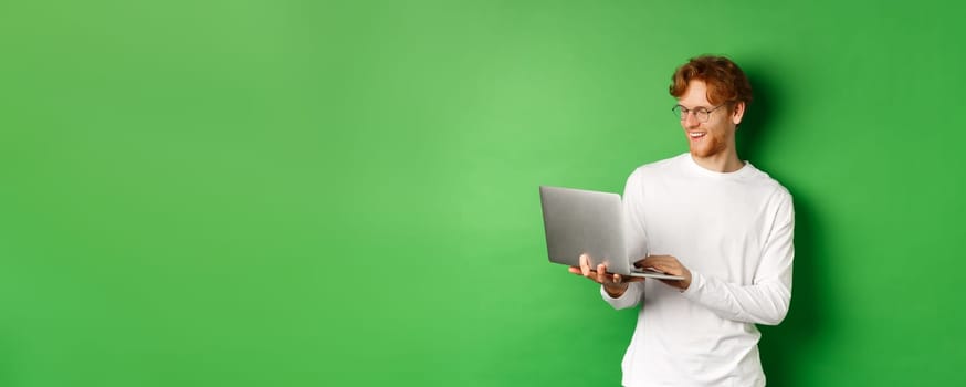 Smiling young man with red hair, wearing glasses, working on laptop and smiling, standing over green background.