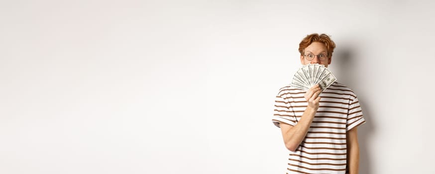 Shopping and finance concept. Lucky redhead guy winning, showing prize money and smiling happy at camera, standing in glasses and t-shirt over white background.