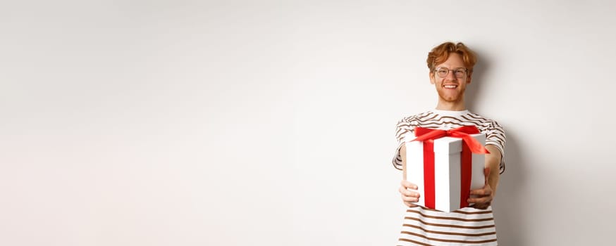 Valentines day and holidays concept. Happy redhead man smiling and giving you gift box, congratulating with birthday, standing over white background.