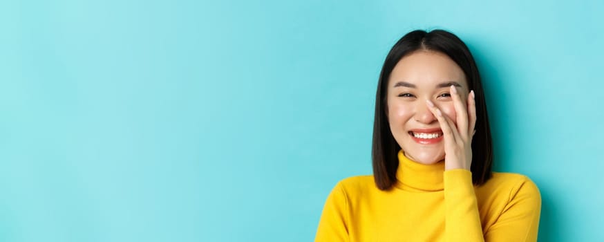 Beauty and makeup concept. Close up of beautiful asian woman blushing and laughing, touching glowing healthy skin, smiling happy, standing over blue background.