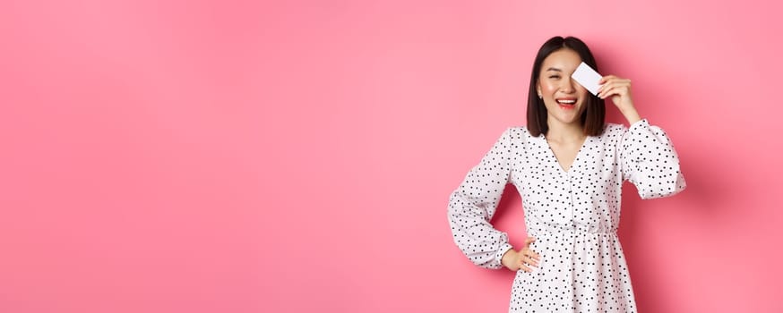 Shopping concept. Pretty asian woman holding bank credit card and smiling, standing over pink background.