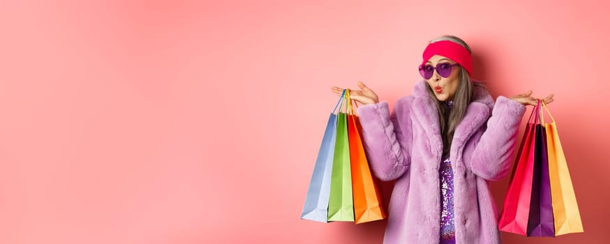 Stylish asian senior woman going shopping, wearing trendy clothes and sunglasses, holding store bags with clothes and purchases, pink background.