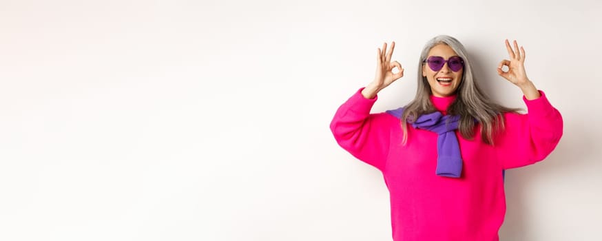 Stylish modern asian grandmother in sunglasses and pink sweater, smiling happy and showing OK signs, approve something cool, white background.
