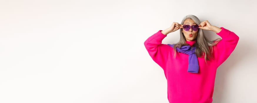 Fashionable asian senior woman in sunglasses and stylish pink sweater pucker lips, raising eyebrows and looking surprised at camera, white background.