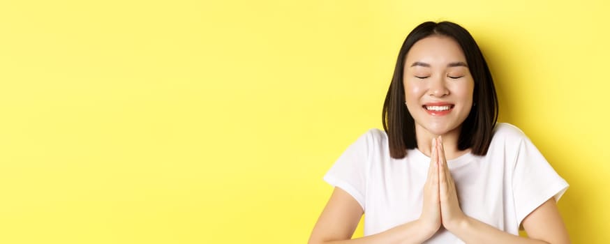 Close up of hopeful asian girl in white t-shirt, holding hands in pray, namste gesture and smiling, making wish or pleading, standing over yellow background.