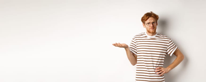 So what. Confused and annoyed redhead man waiting for reply, standing questioned with raised hand, holding something on palm, white background.