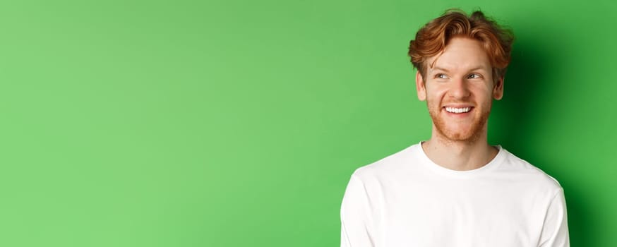 Headshot of happy redhead man with beard wearing white long sleeve, looking left at copy space and smiling, standing over green background.