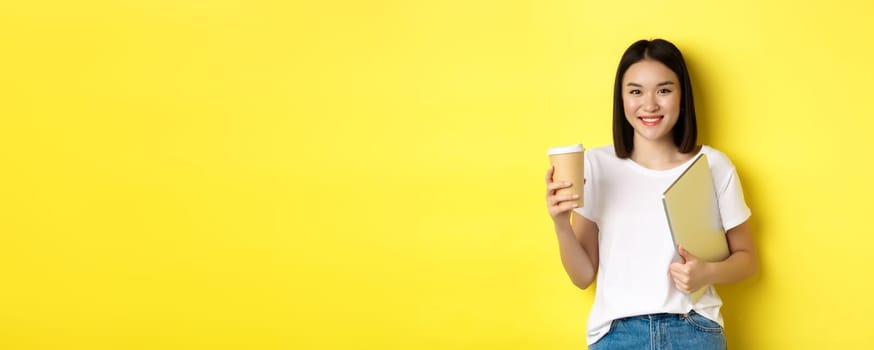 Asian girl student drinking coffee and holding laptop, smiling at camera, standing over yellow background.