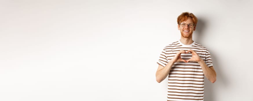 Valentines day. Happy boyfriend with red hair, smiling and showing heart gesture, I love you, standing over white background.