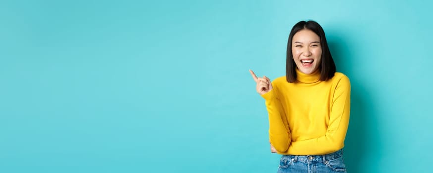 Image of carefree pretty korean girl laughing and looking happy, pointing finger at upper left corner promo, standing against blue background.