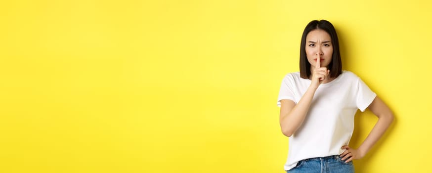 Beauty and fashion concept. Angry and bossy asian girl telling to be quiet, scolding loud people, shushing and frowning at camera, standing in t-shirt over yellow background.
