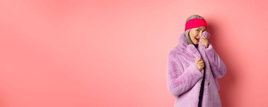 Stylish asian elderly woman hiding face behind coat collar, smiling and looking at camera silly. Modern grandmother posing against white background.